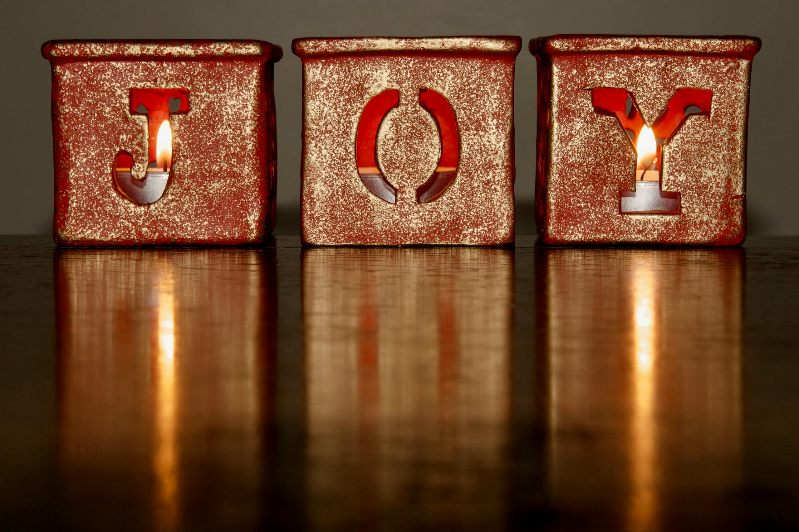 Three candlelit blocks spelling 'JOY' with a glowing warm reflection on a wooden table.